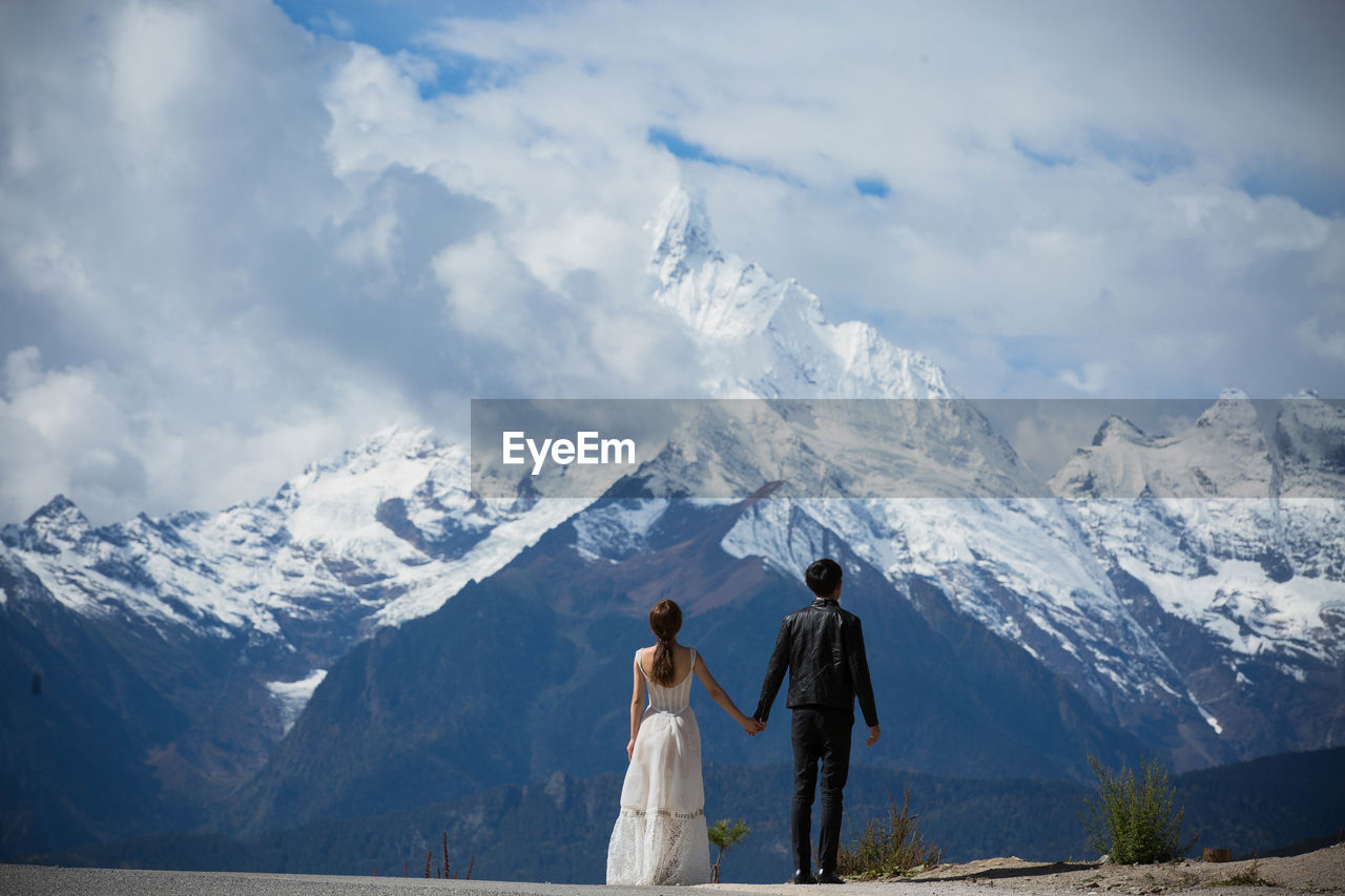 Couple standing in front of snowcapped mountains against cloudy sky