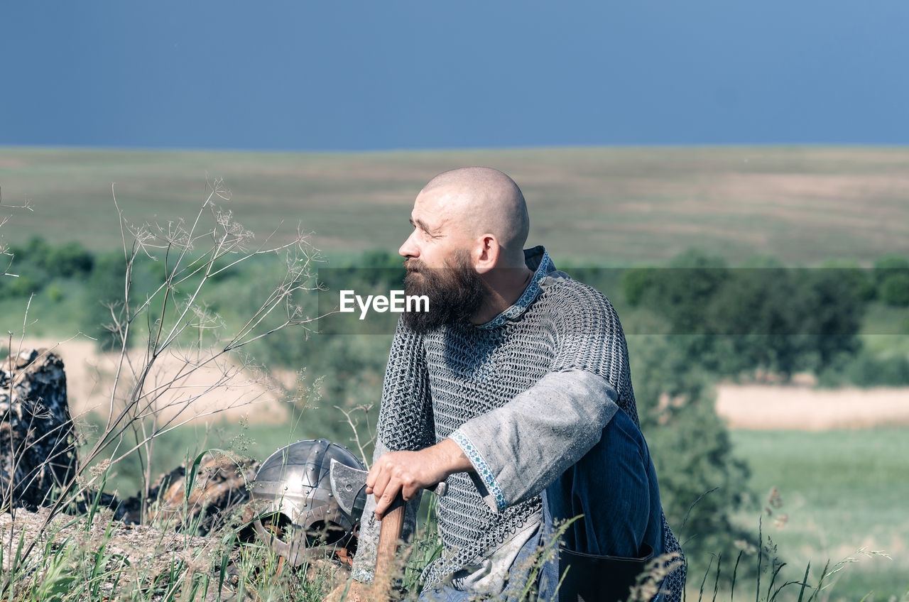 Viking. bald bearded man in metal chain mail over linen shirt sits on hill, leaning on ax. 