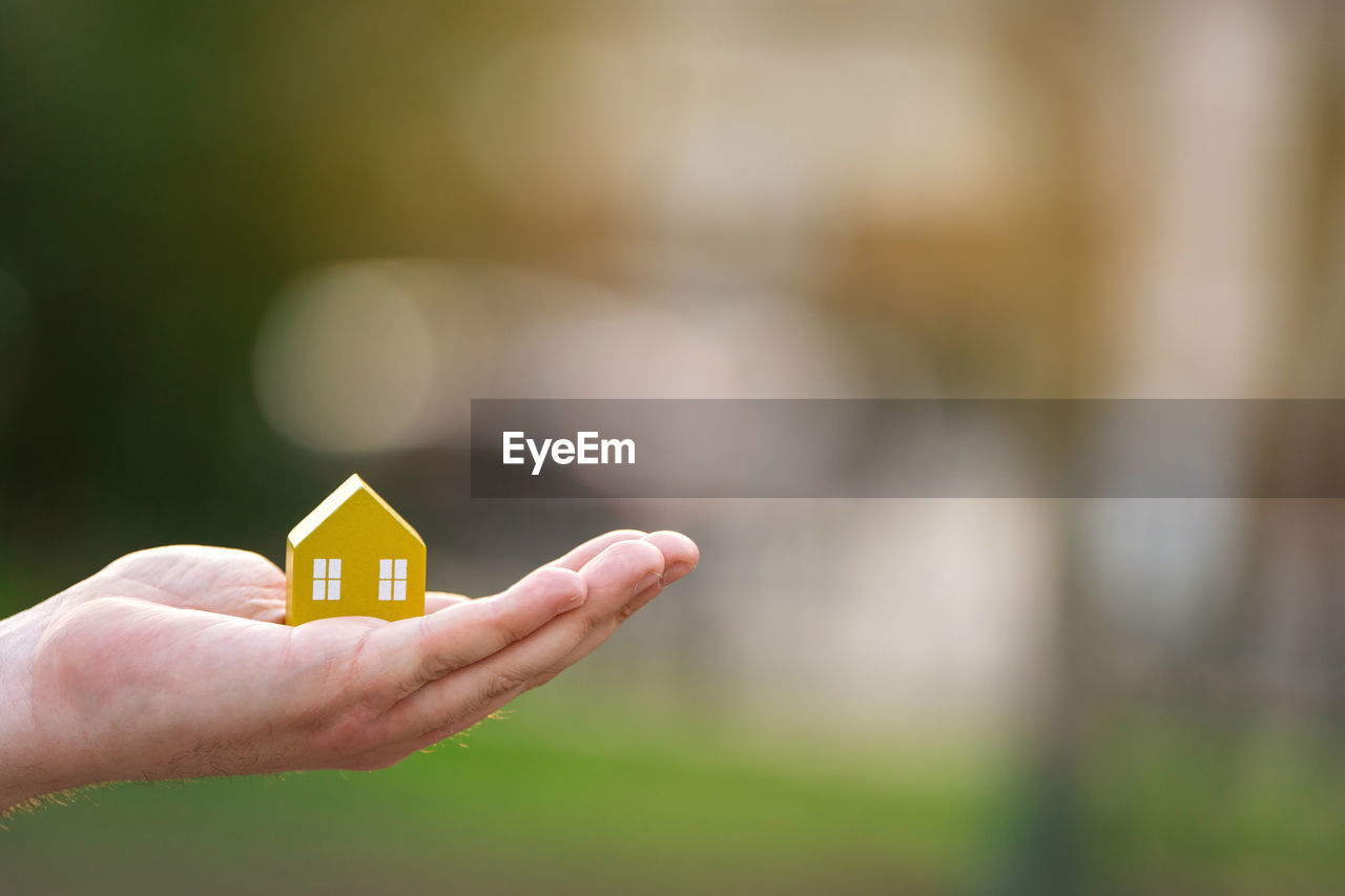 A man holds a yellow house on his hand outdoors. 