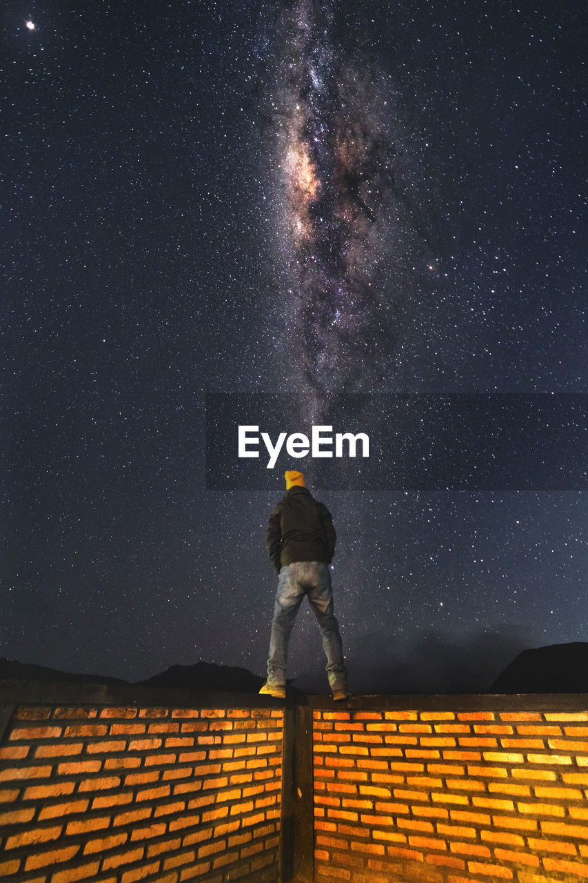 Rear view of man standing on brick wall against star field at night