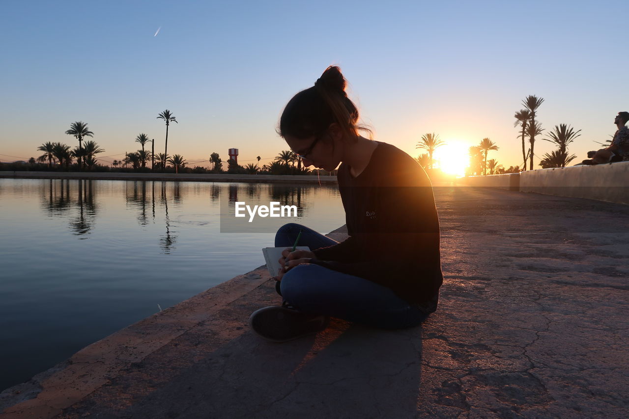 Full length of woman writing on diary while sitting by lake