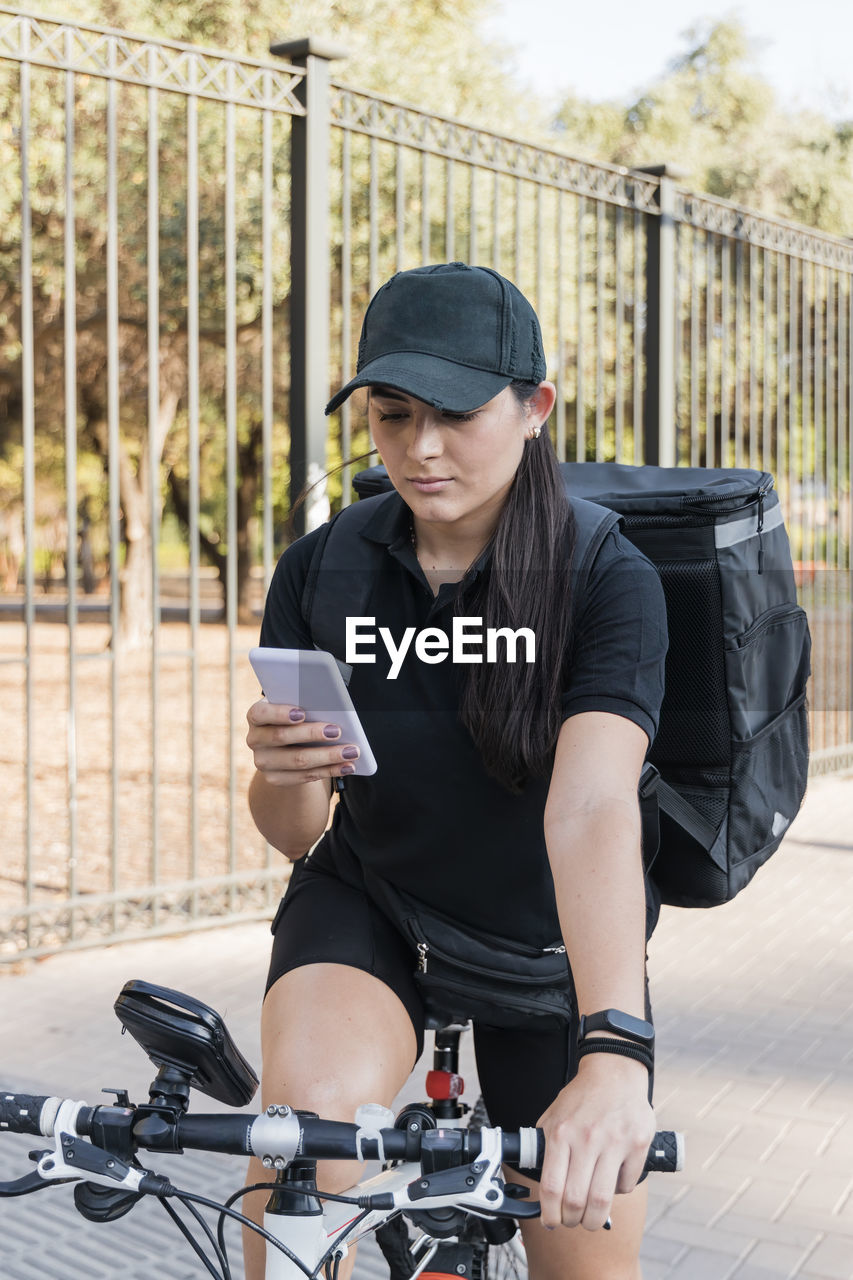 Young delivery female sitting on bicycle while using smart phone