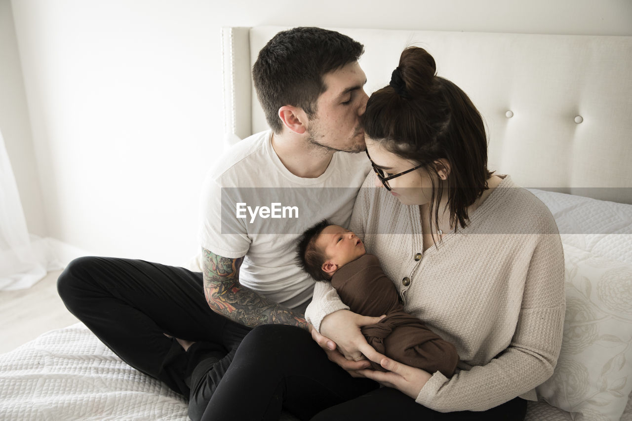 Tattooed millennial dad kisses mom while she holds their newborn