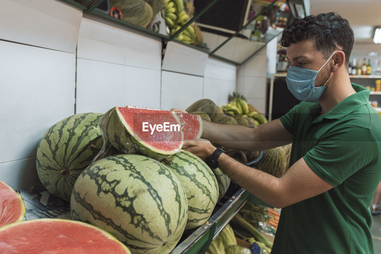 A young man wearing a mask placing watermelons in the fruit shop.