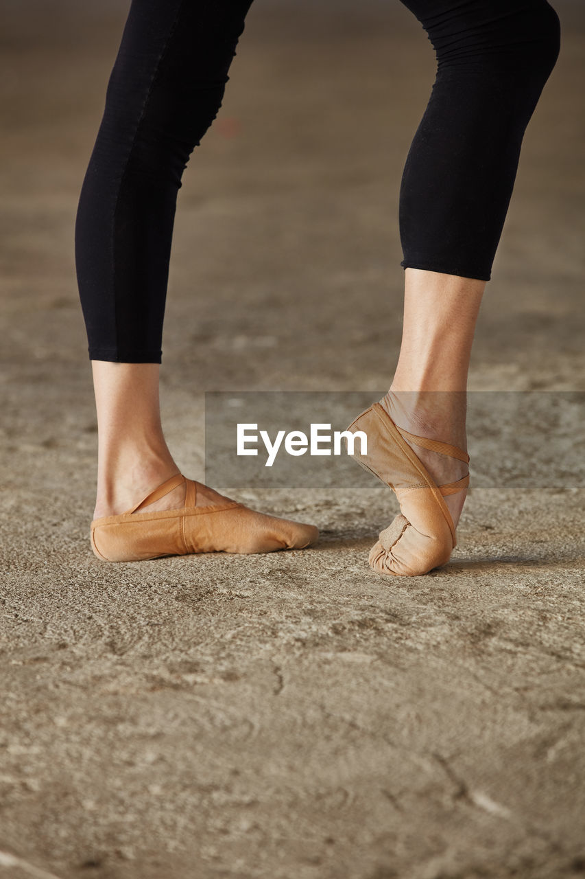 Crop faceless ballet dancer in black leotard and pointe shoes stretching legs with pose on concrete floor