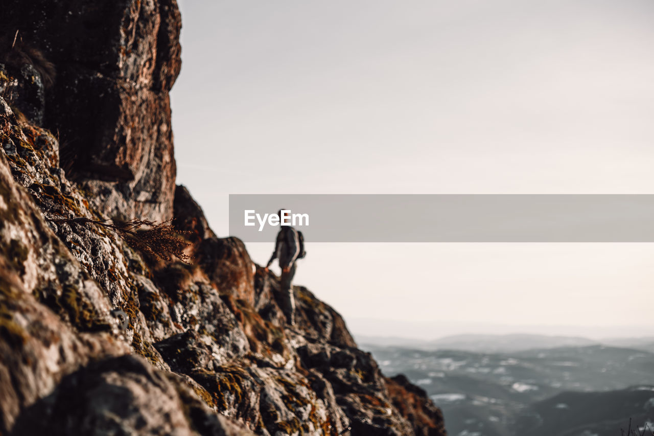 REAR VIEW OF MAN ON ROCK BY MOUNTAIN AGAINST SKY