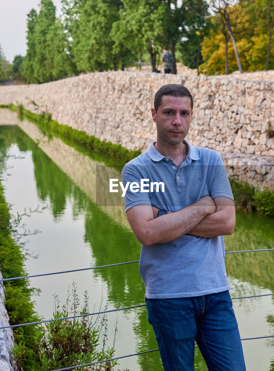 one person, water, men, adult, standing, portrait, looking at camera, plant, nature, front view, casual clothing, three quarter length, arms crossed, lake, tree, day, person, clothing, reflection, outdoors, beard, occupation, lifestyles, facial hair, smiling, emotion, jeans, leisure activity, mature adult