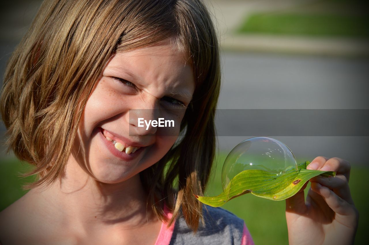 Close-up portrait of smiling girl holding bubble on leaf during sunny day