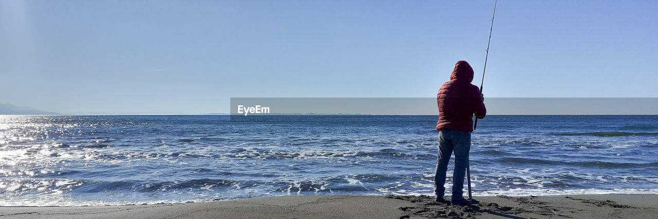 REAR VIEW OF MAN STANDING ON BEACH AGAINST SKY