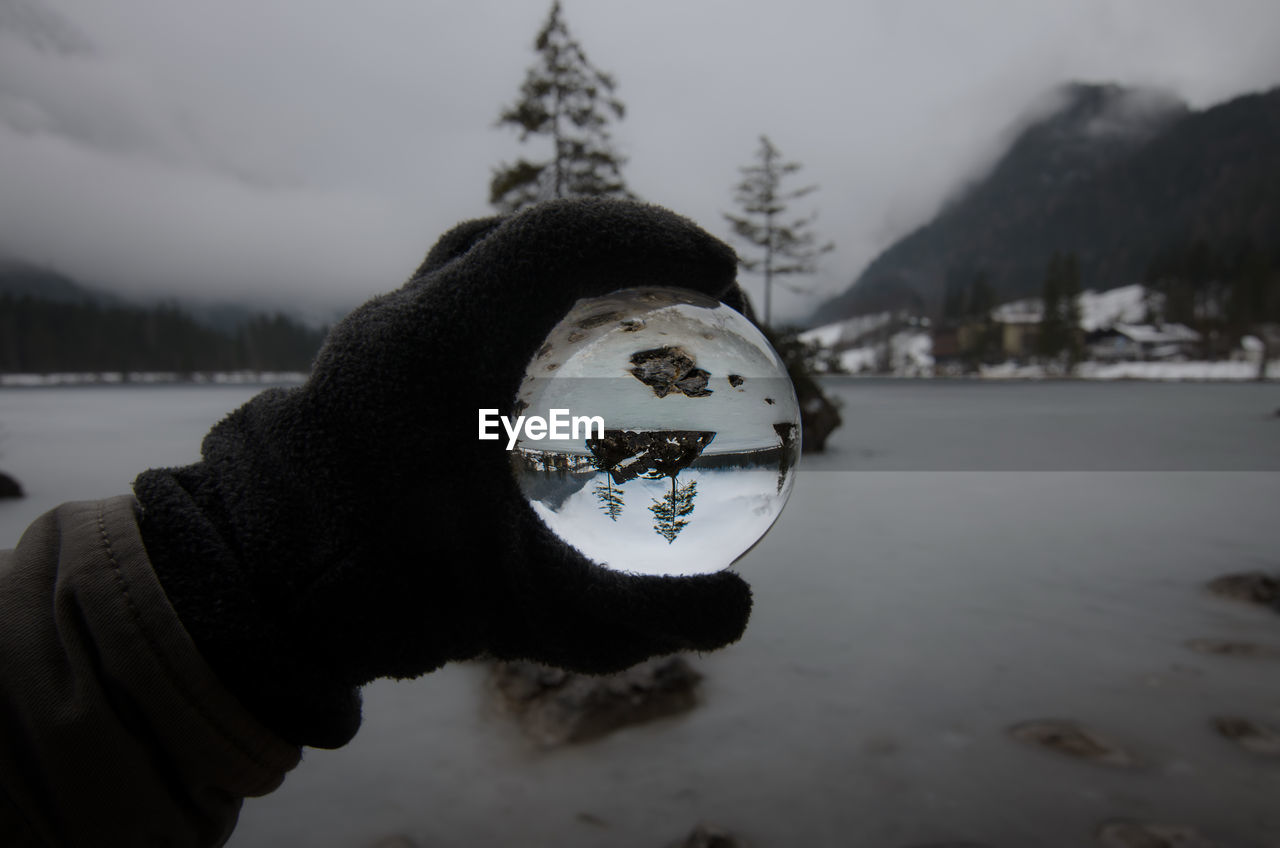Cropped hand holding crystal ball by lake reflection during winter