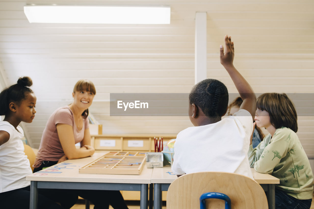 Teacher looking at boy raising hand while answering in classroom