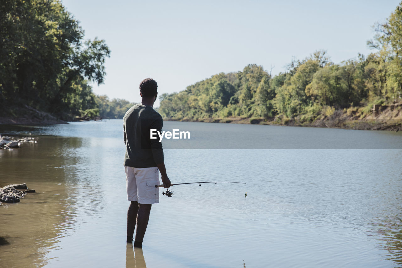 Rear view of man holding fishing rod while standing in lake against clear sky