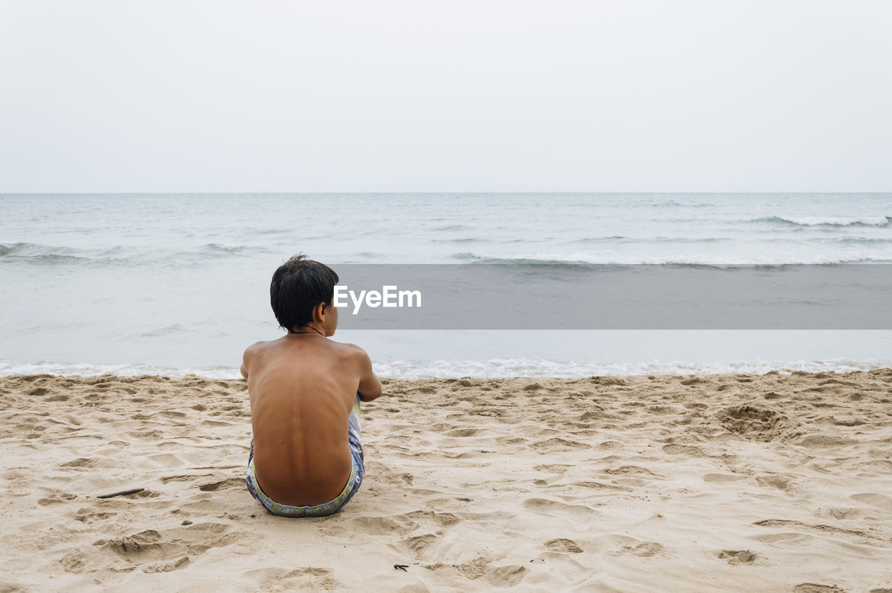 Rear view of shirtless young man sitting at beach against clear sky