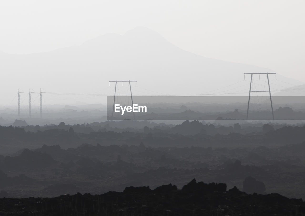 Black and white of power lines with wires placed on ground with rough surface against mountain range covered with fog