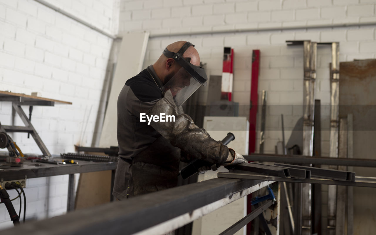 Serious male worker in dirty apron standing at workbench and preparing metal details for welding