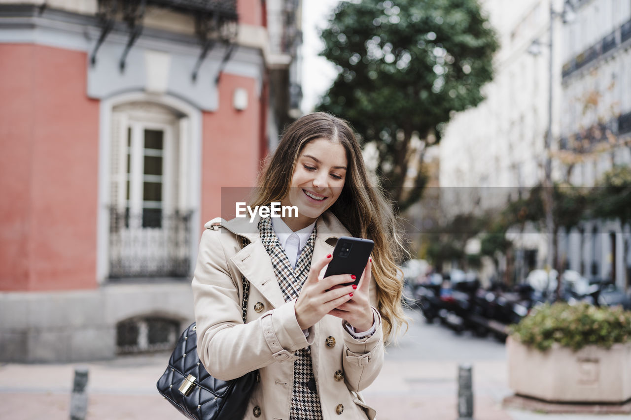 Happy woman carrying purse while using mobile phone in city