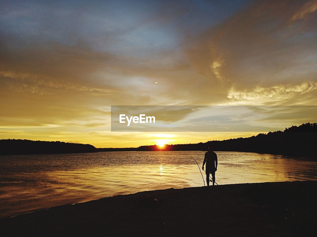 Silhouette man standing by lake against sky during sunset