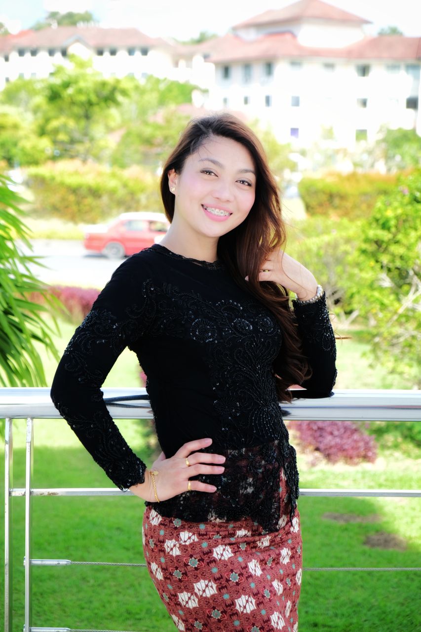 PORTRAIT OF SMILING YOUNG WOMAN STANDING AGAINST WALL