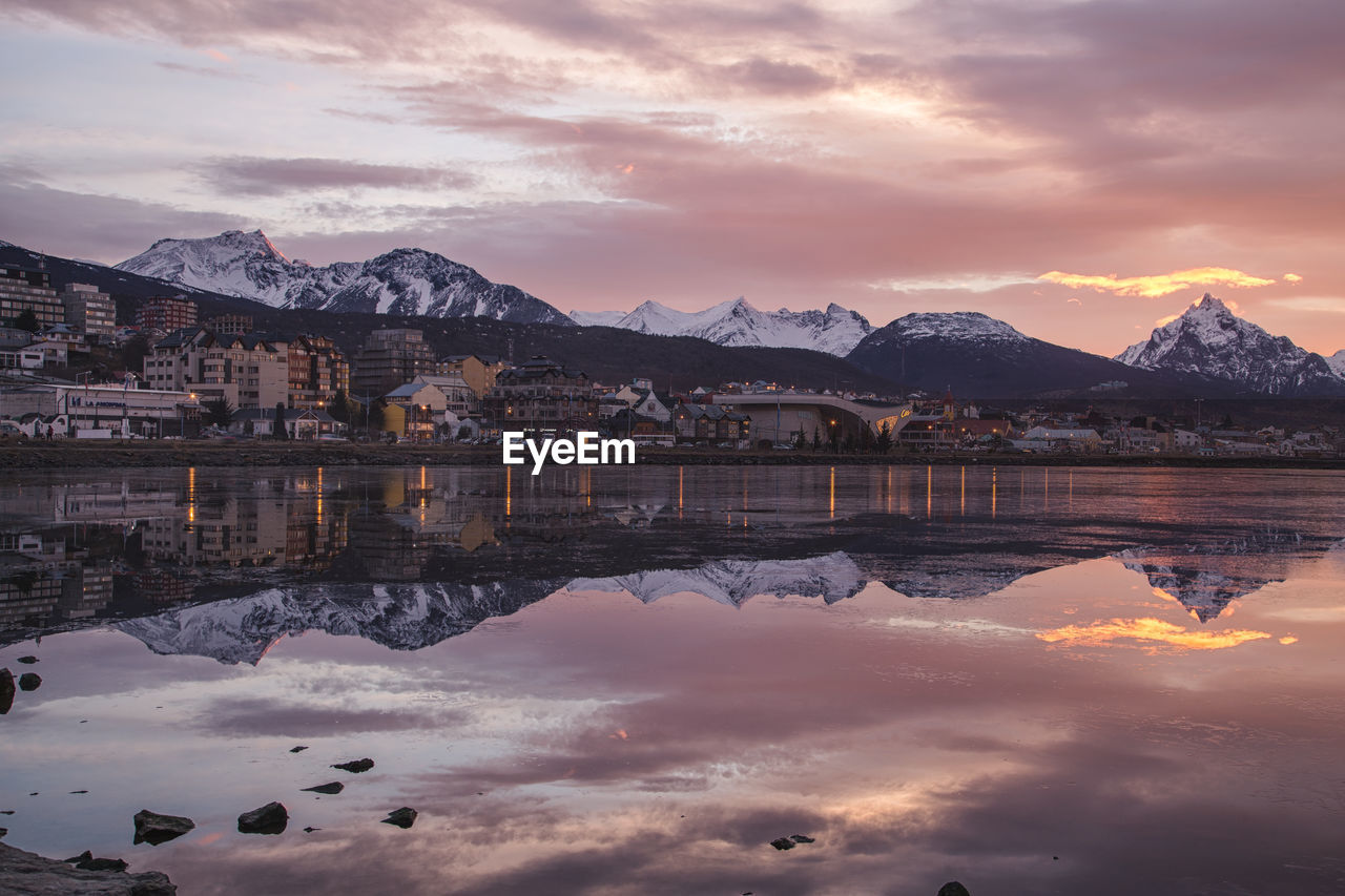 Reflection of buildings and snowcapped mountains in lake during sunset