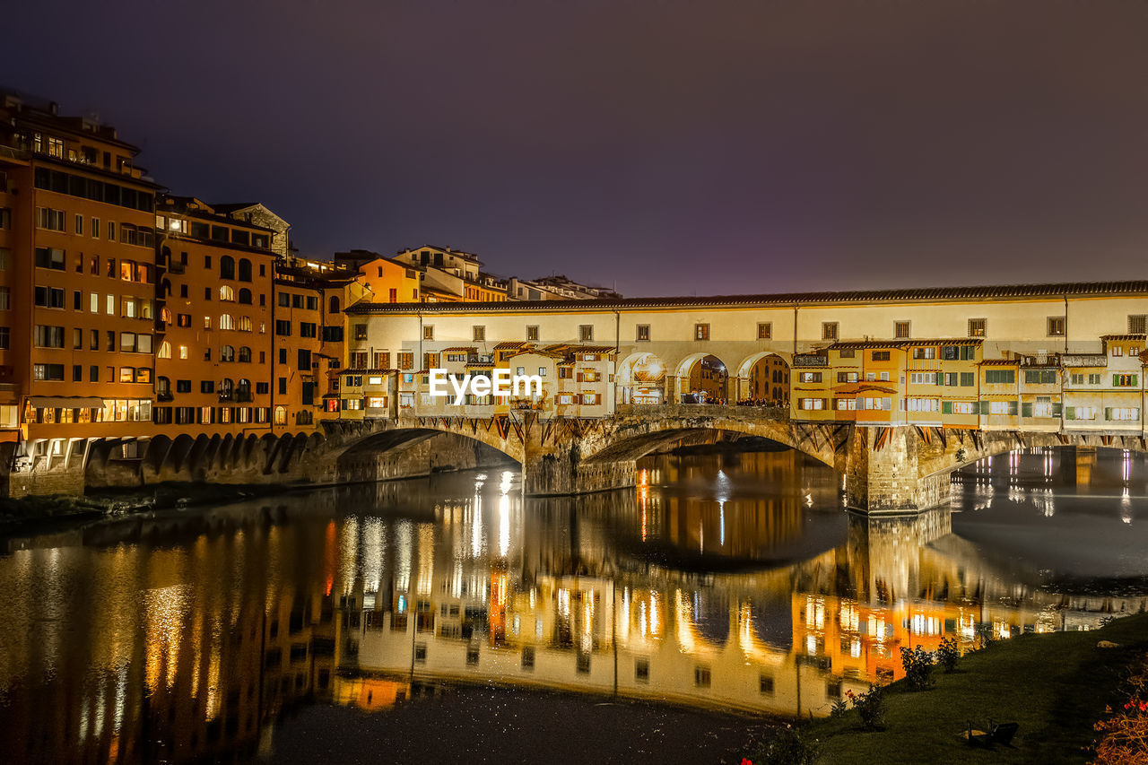 BRIDGE OVER RIVER BY ILLUMINATED BUILDINGS AT NIGHT