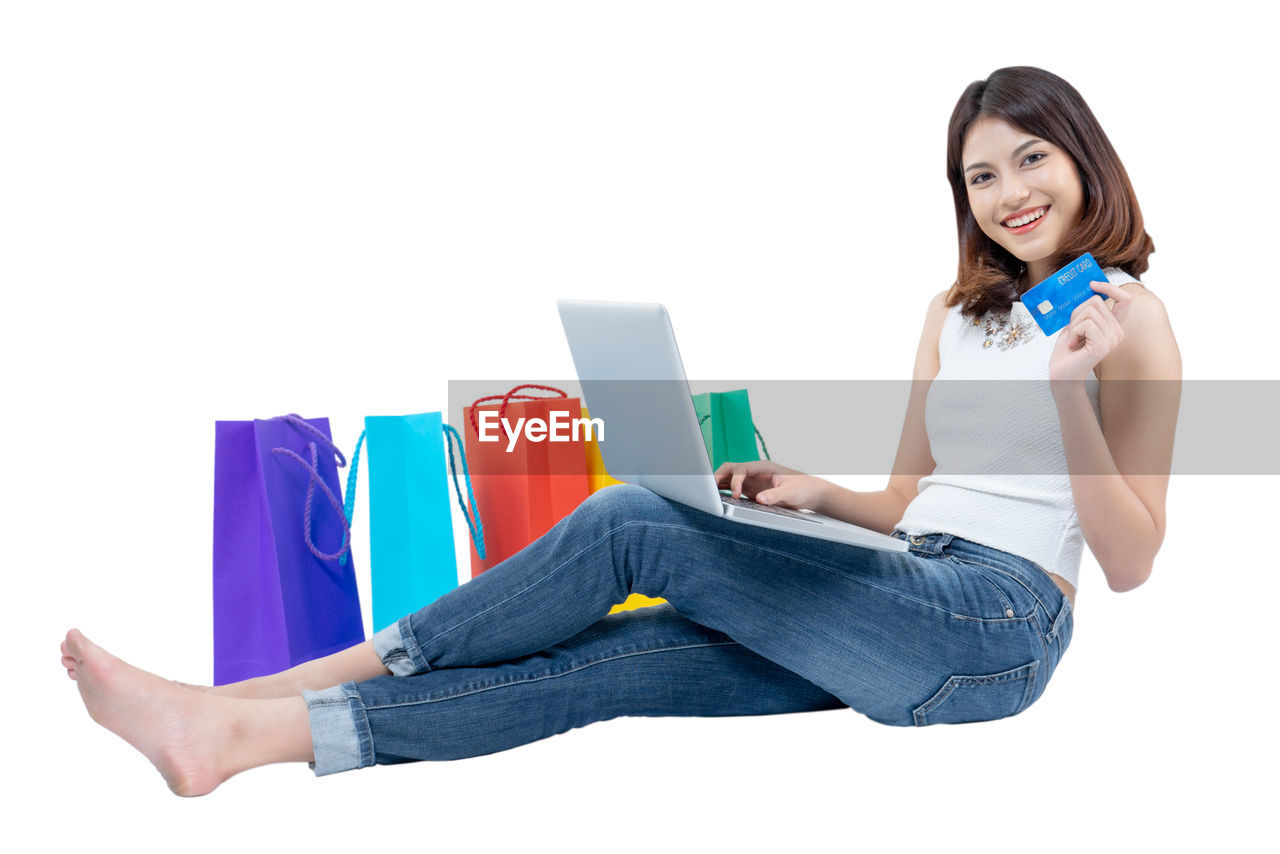 Smiling young woman showing credit card while using laptop by colorful paper bags against white background