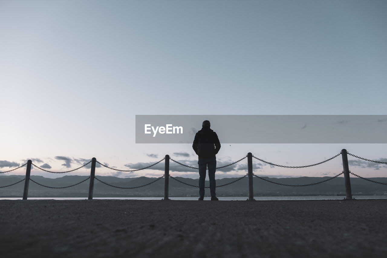 REAR VIEW OF MAN STANDING ON BEACH AGAINST CLEAR SKY