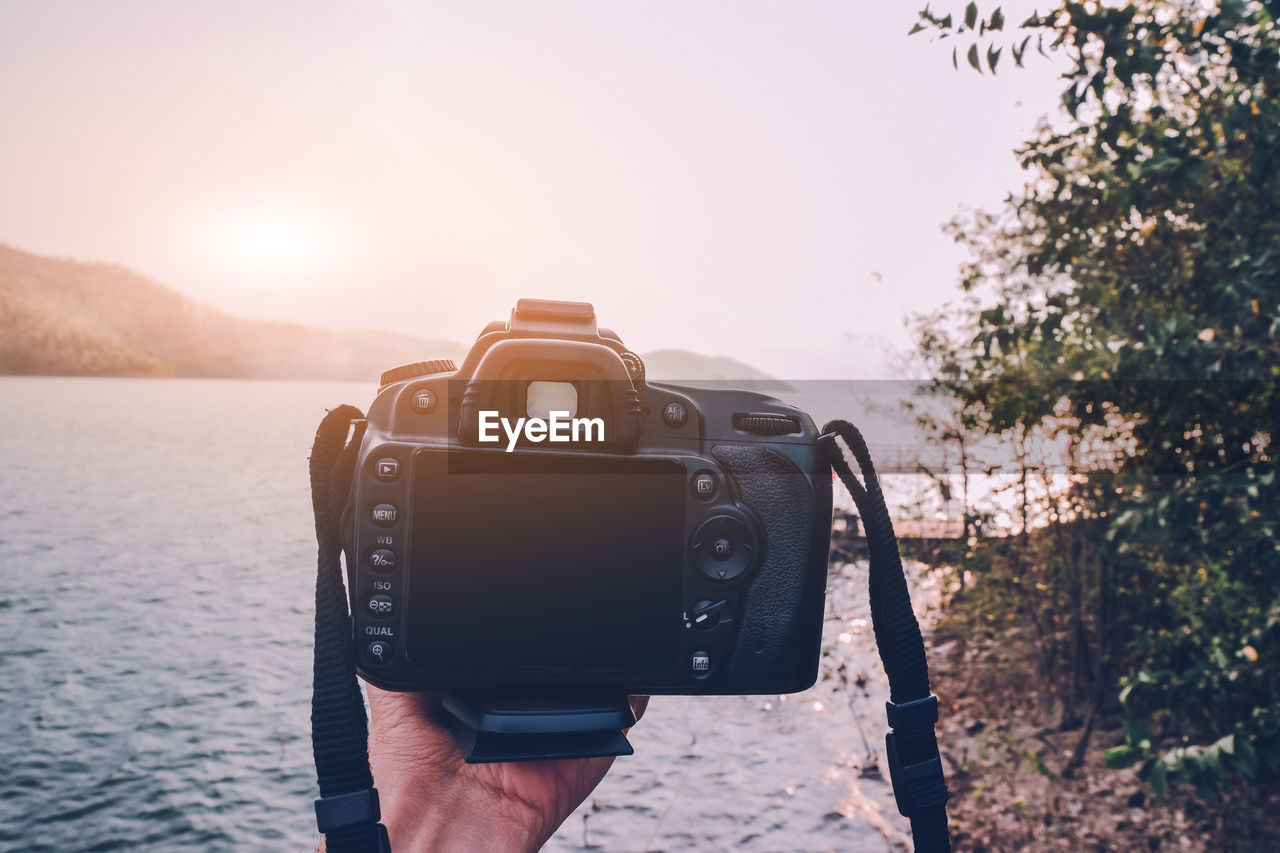 Cropped hand holding camera by lake during sunset