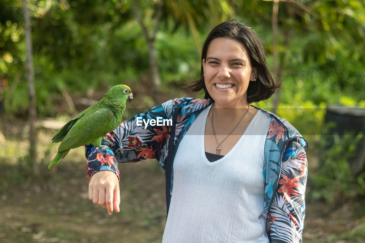 Woman poses with a smile as she carries her arm to a macaw in a camp