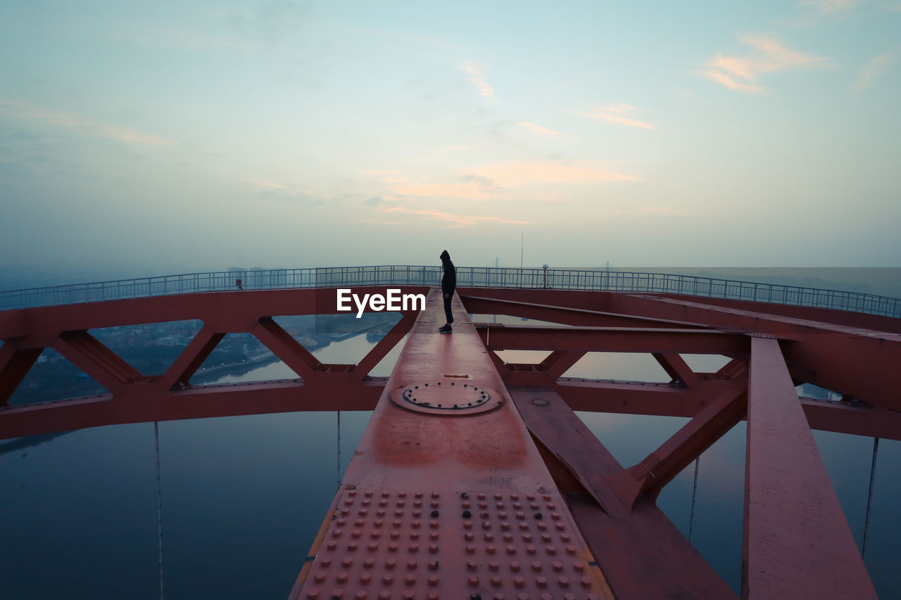 Man standing on railing over river against sky