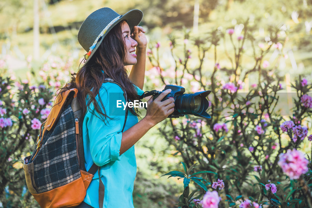 Side view of smiling young woman holding camera while standing amidst flowers on land