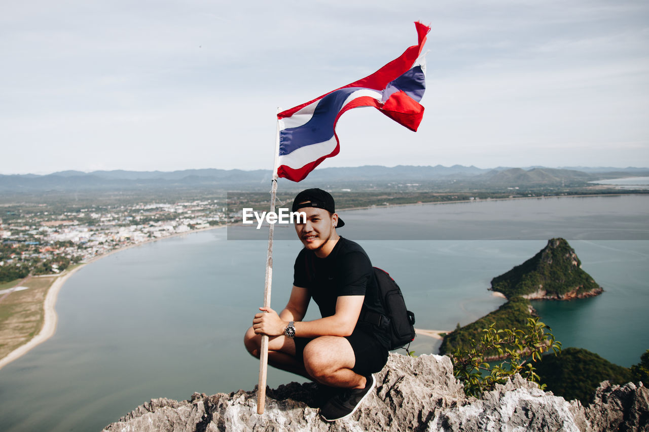 Portrait of smiling young man holding flag while crouching on mountain against sea at beach