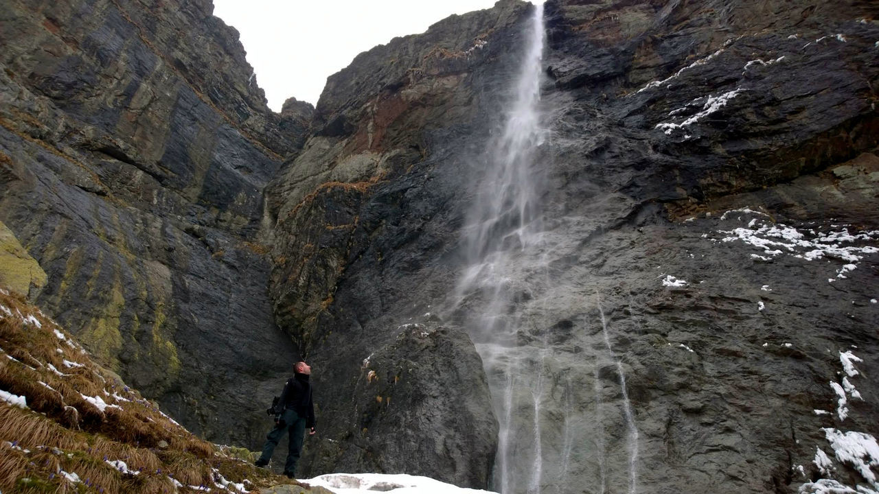 REAR VIEW OF MAN STANDING BY WATERFALL AGAINST MOUNTAIN