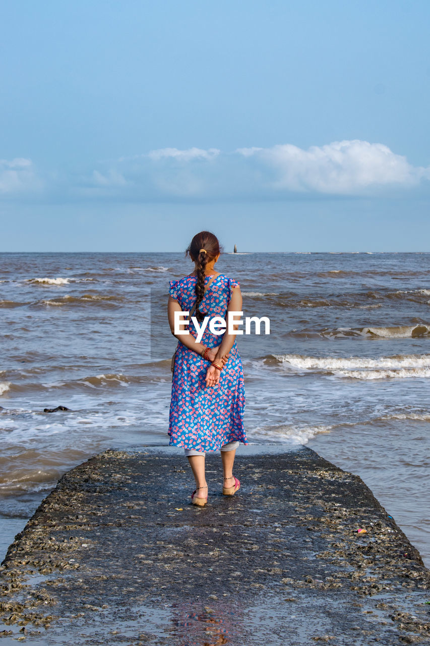 water, sea, beach, land, one person, sky, blue, shore, full length, ocean, vacation, coast, nature, standing, body of water, horizon over water, horizon, leisure activity, adult, casual clothing, women, wave, beauty in nature, holiday, day, trip, front view, cloud, clothing, young adult, rock, lifestyles, dress, outdoors, scenics - nature, looking, fashion, tranquility, child, female, sand, person, emotion, motion, childhood