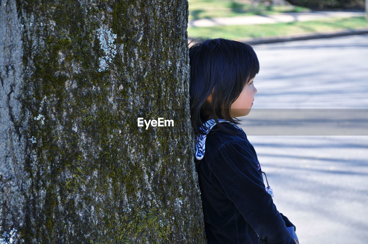 Side view of girl leaning against a tree trunk