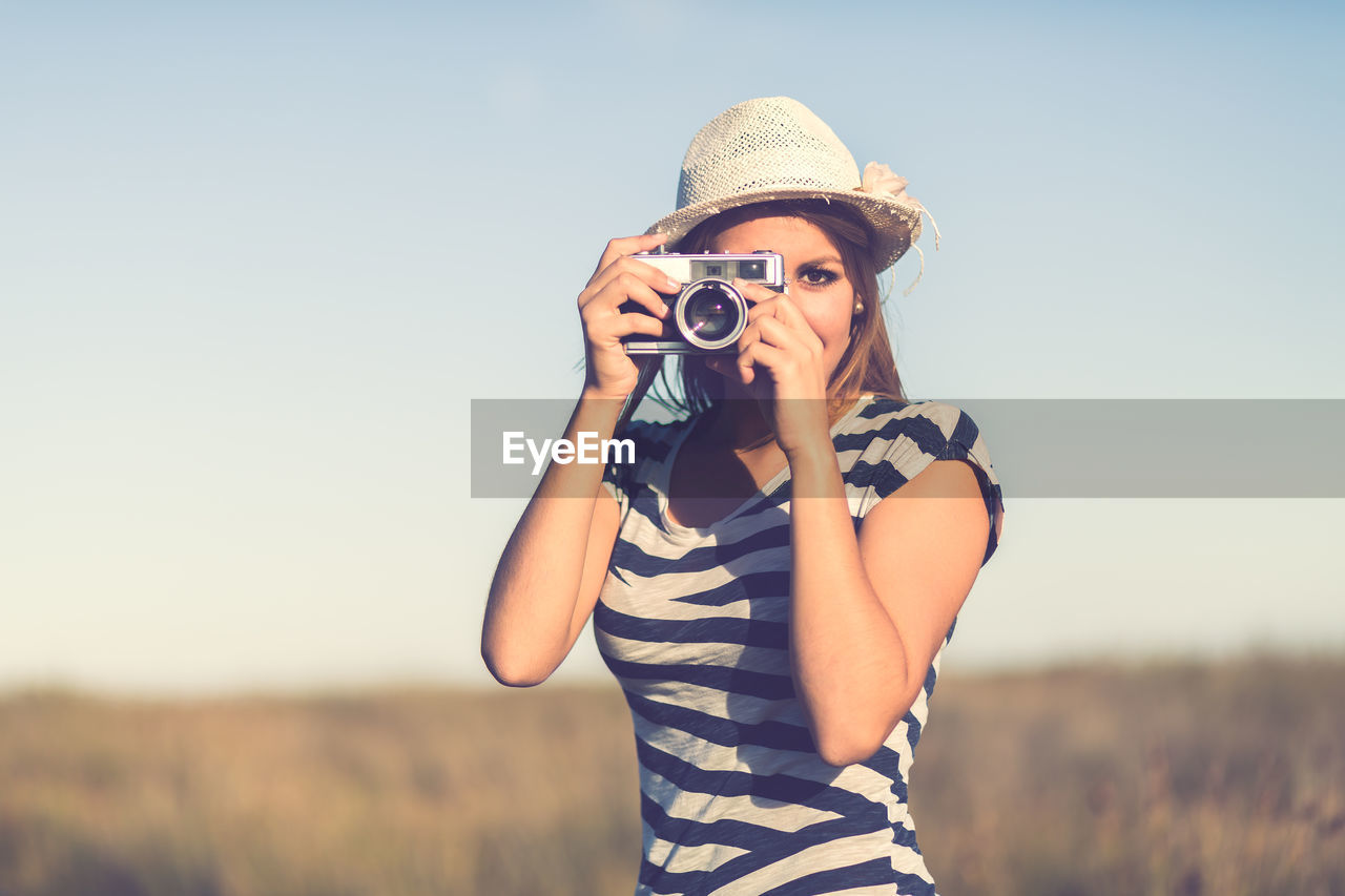 Young woman photographing with camera on field against clear sky