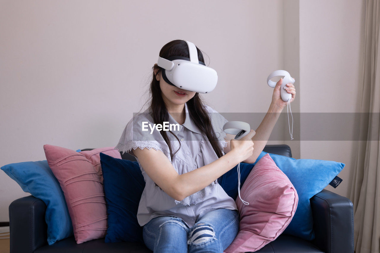 indoors, one person, sitting, adult, domestic room, furniture, women, blue, sofa, lifestyles, domestic life, relaxation, casual clothing, holding, young adult, technology, living room, person, home interior, clothing, limb, three quarter length, full length, pillow, female, bed, front view, leisure activity, smiling, enjoyment, happiness, human leg