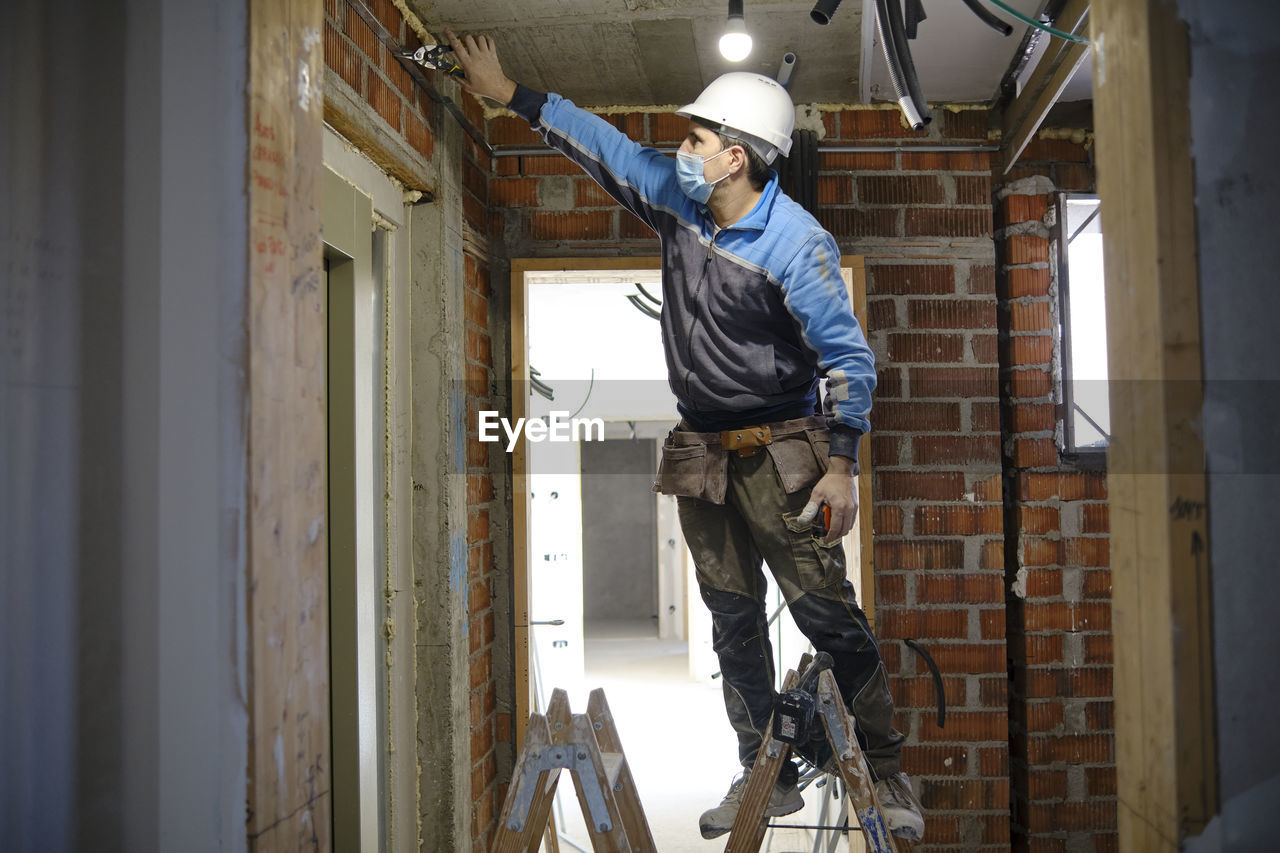 Electrician working with tool standing on ladder at construction site