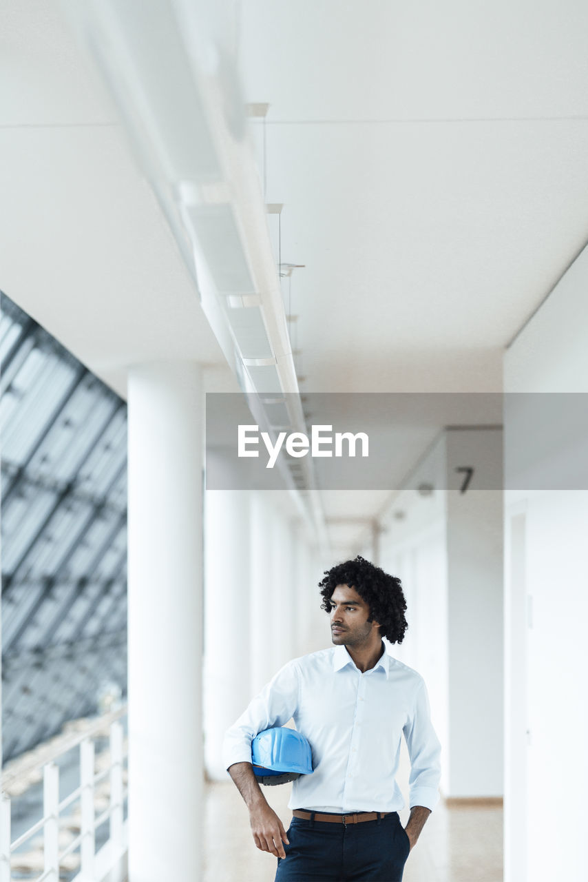 Young male engineer with hardhat looking away while standing in corridor at office