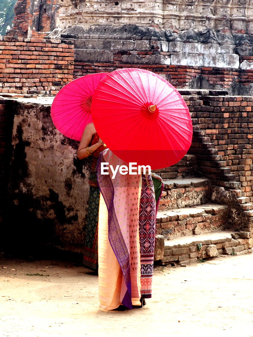 WOMAN WITH UMBRELLA STANDING AGAINST BRICK WALL DURING RAINY SEASON