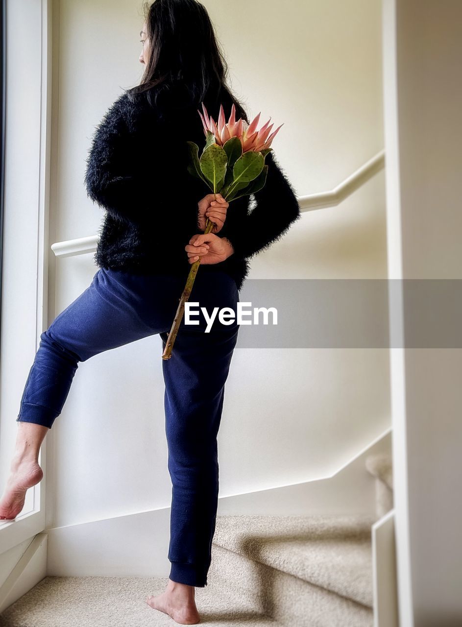 Low section of woman standing against wall and holding king protea flower