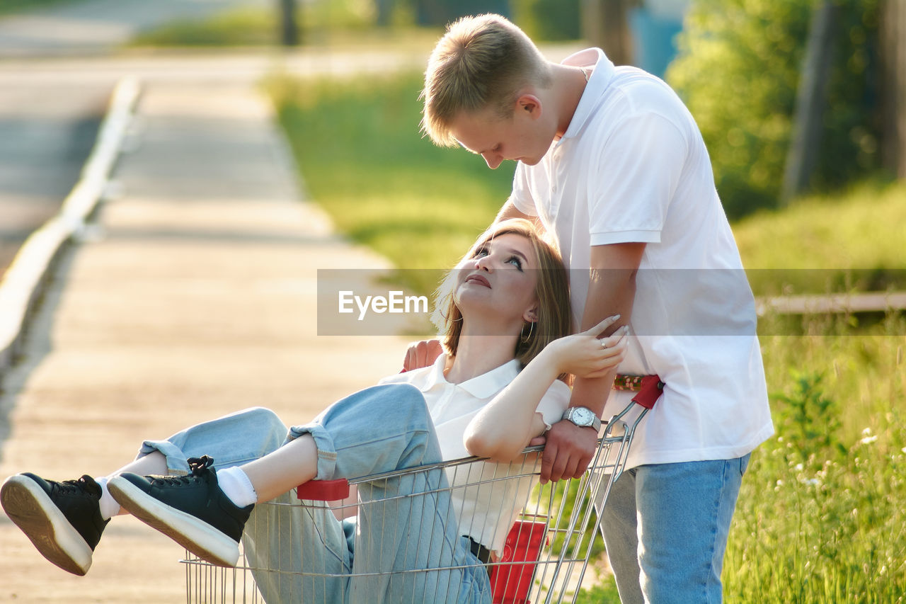 A couple is having fun, a guy is pushing a shopping cart, and his girlfriend is sitting there.