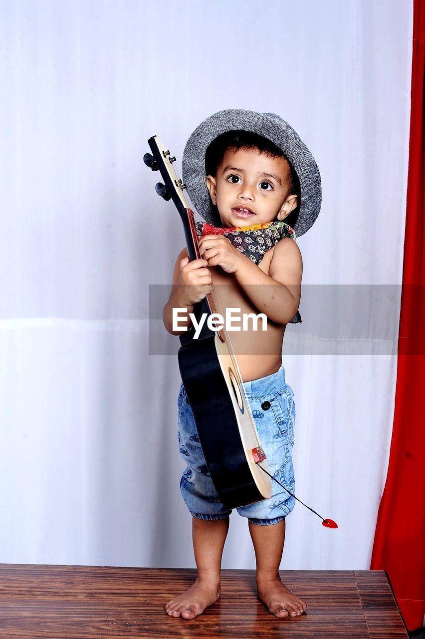 Full length portrait of cute boy standing against wall wearing a hat