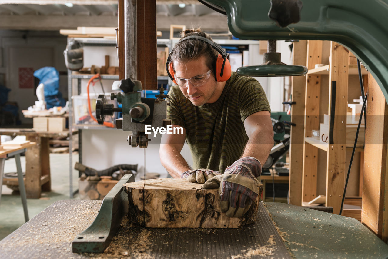 Focused male carpenter in protective ear muffs and goggles cutting piece of wood with band saw in shabby workshop