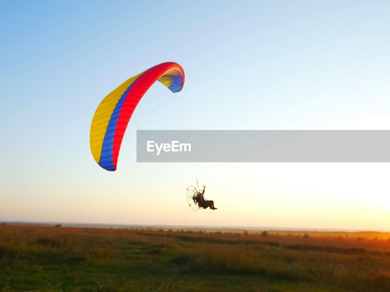 Man flying powered parachute over grassy field against clear sky during sunset