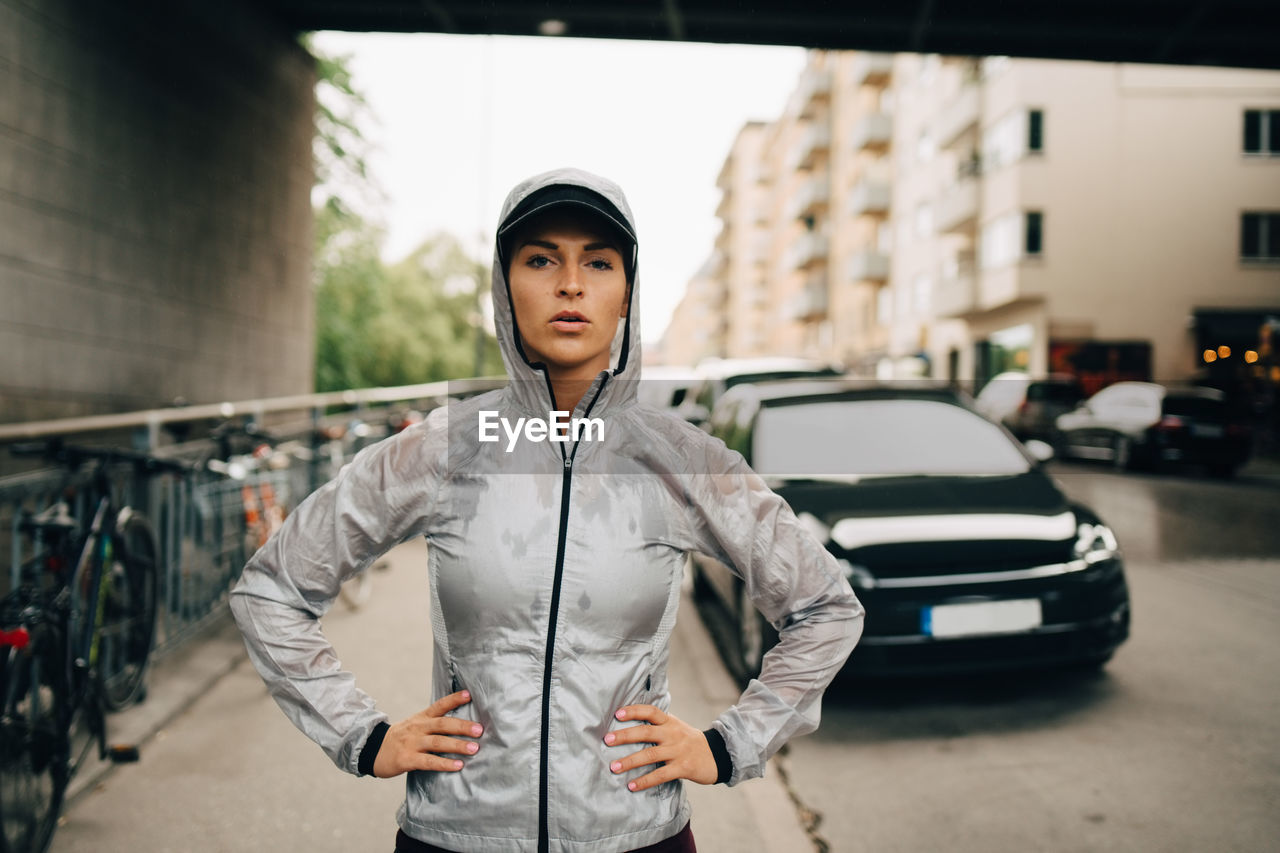 Portrait of confident female athlete in wet hooded jacket standing on sidewalk in city