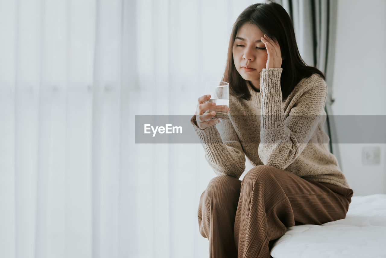 Woman with headache holding glass while sitting on bed at home