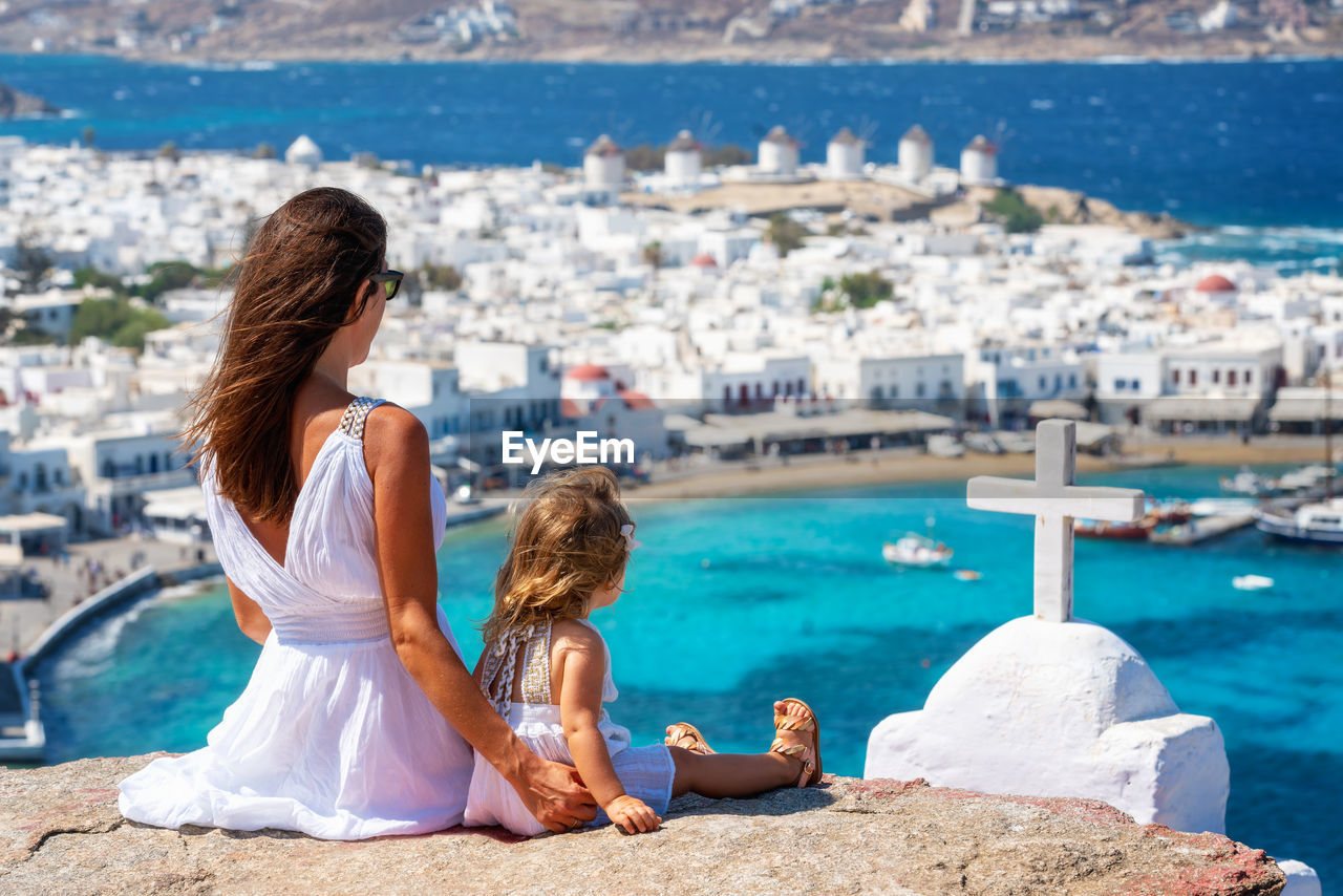 Rear view of mother and daughter sitting on cliff against townscape