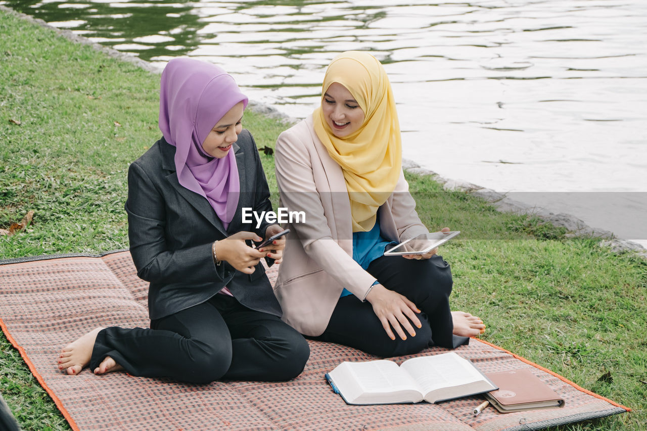 High angle view of female friends using wireless technology while sitting on mat by lake