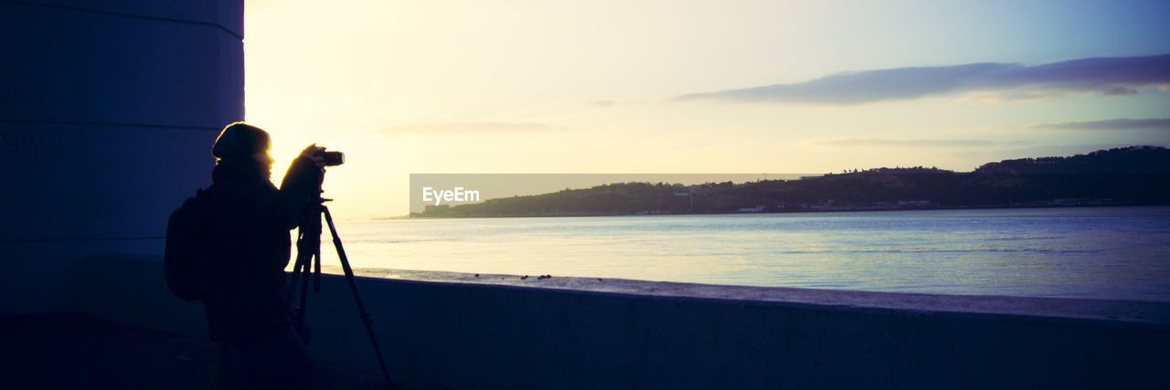 Panoramic shot of silhouette woman with photographing at river shore