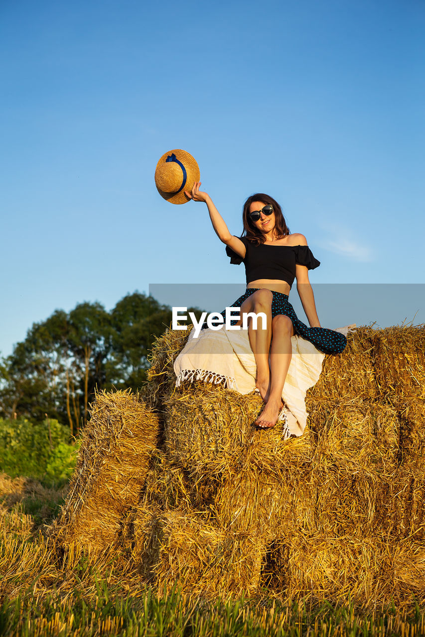A young woman looks at the setting sun and holds a straw hat, sits on a pile of straw bales.
